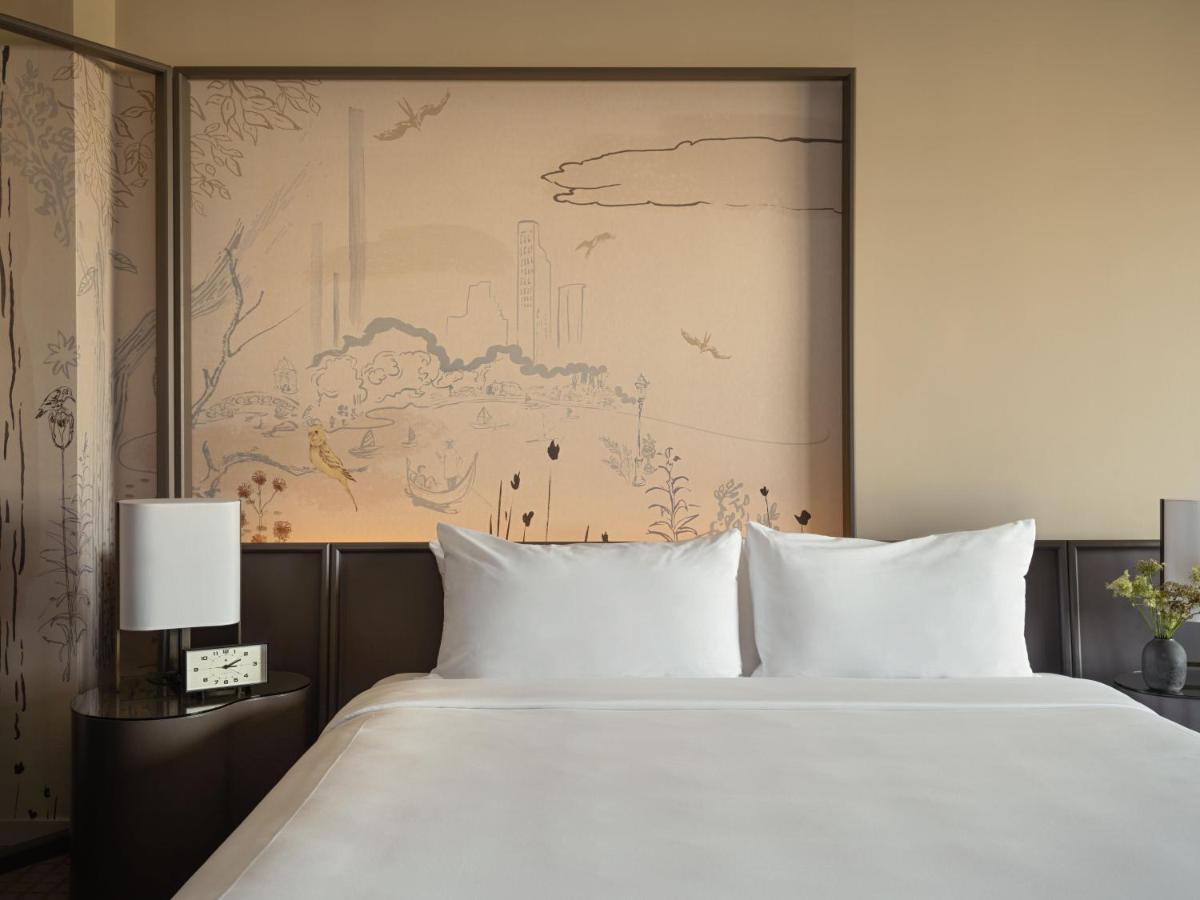 Park Lane New York酒店 外观 照片 The photo depicts a modern hotel room featuring a plush bed with white linens and two pillows. Behind the bed is an artistic wall mural that appears to illustrate a serene landscape with subtle outlines of trees and clouds. On either side of the bed,