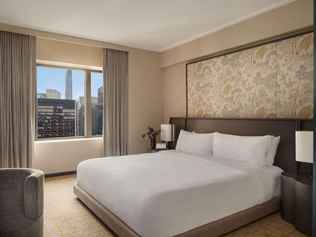 Park Lane New York酒店 客房 照片 The photo shows a modern hotel room featuring a large bed with white bedding against a decorative headboard. There are two bedside lamps on either side of the bed. The room has light-colored walls and soft carpeting. A large window allows natural lig