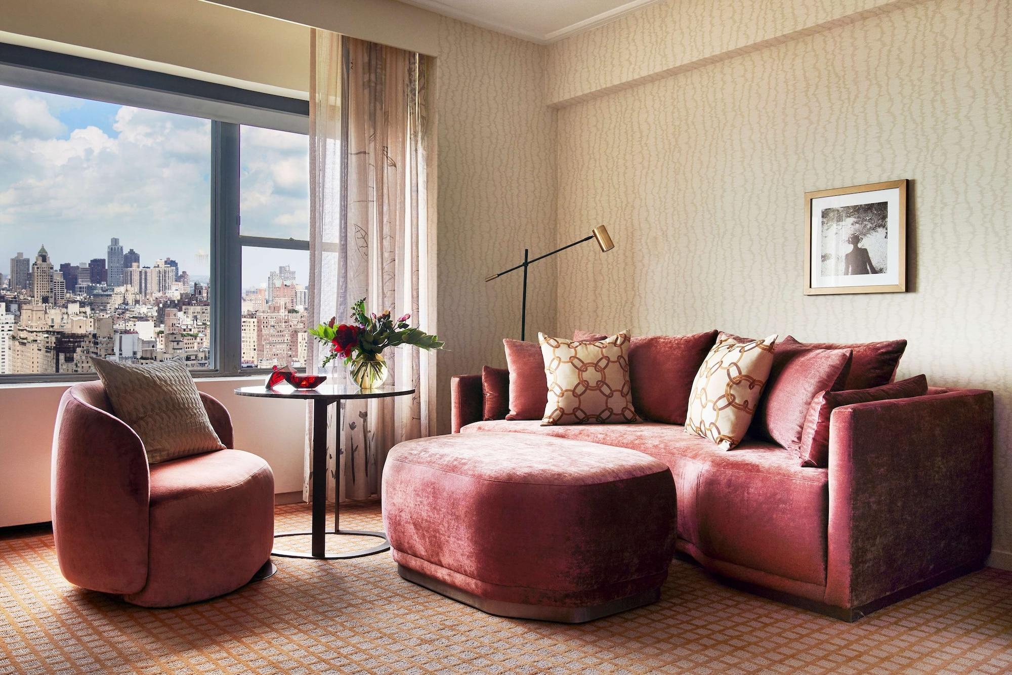 Park Lane New York酒店 外观 照片 The photo shows a modern indoor living space, likely a hotel room or a stylish apartment. There is a plush, curved sectional sofa in a soft pink hue, adorned with decorative throw pillows. A round table sits in front of the sofa, accompanied by a sin