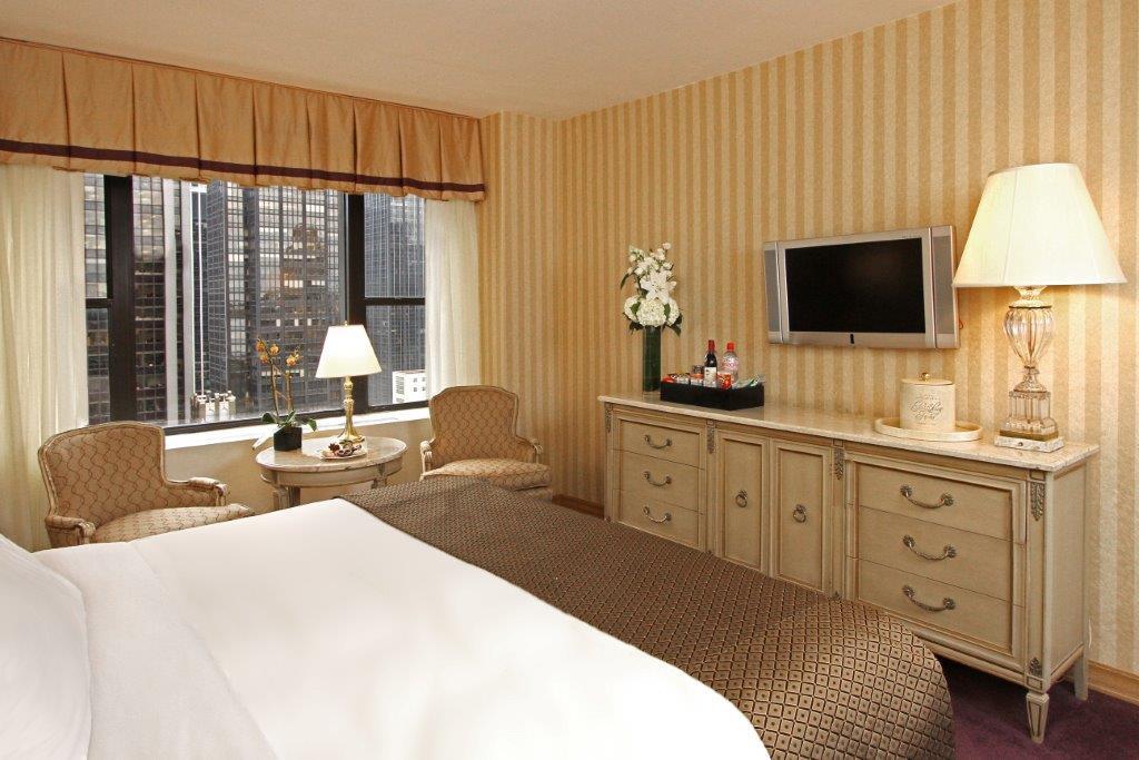 Park Lane New York酒店 外观 照片 The image shows a hotel room with a cozy and inviting atmosphere. There is a large bed with white bedding, and the walls are decorated with striped wallpaper in soft colors. A pair of armchairs sits next to a small table near a window, which allows n