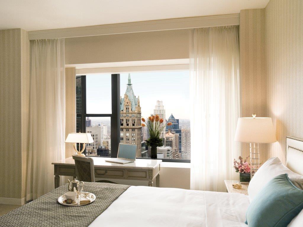 Park Lane New York酒店 外观 照片 The photo depicts a stylish hotel room with a neatly made bed featuring white linens and a decorative throw. There's a modern desk near a large window, which offers a view of a city skyline with notable buildings. The room features soft color tones a